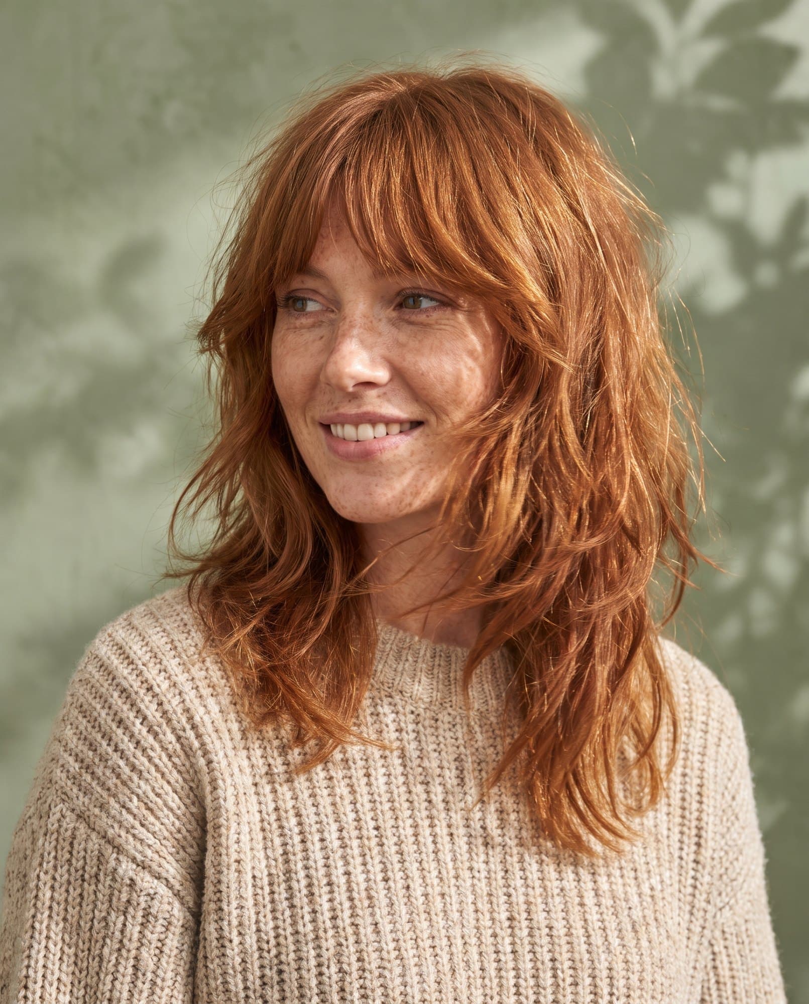 A smiling woman with a textured auburn shag and curtain fringe in a knit jumper.