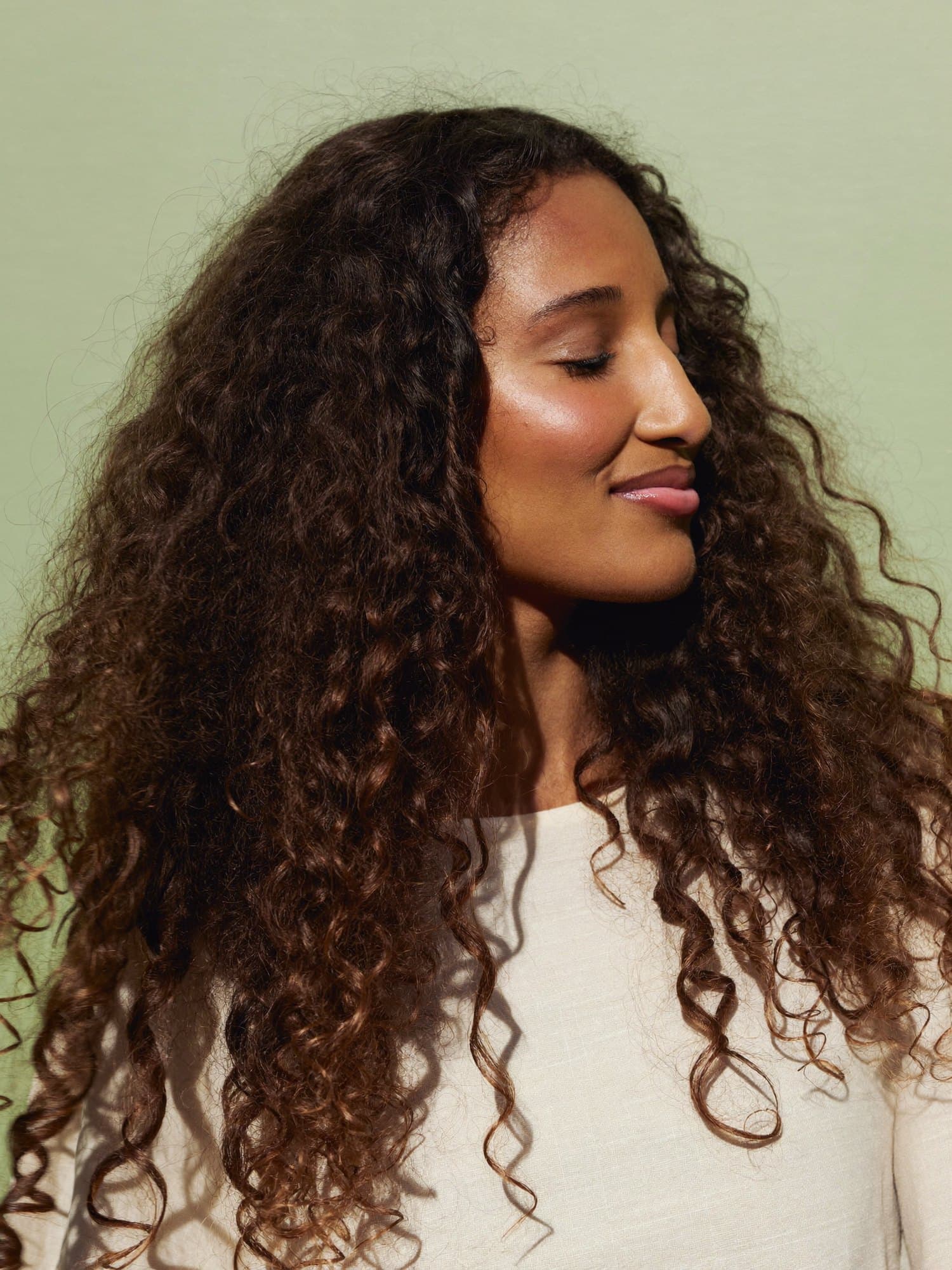 A woman with long, defined natural spiral curls, eyes closed in the sun.