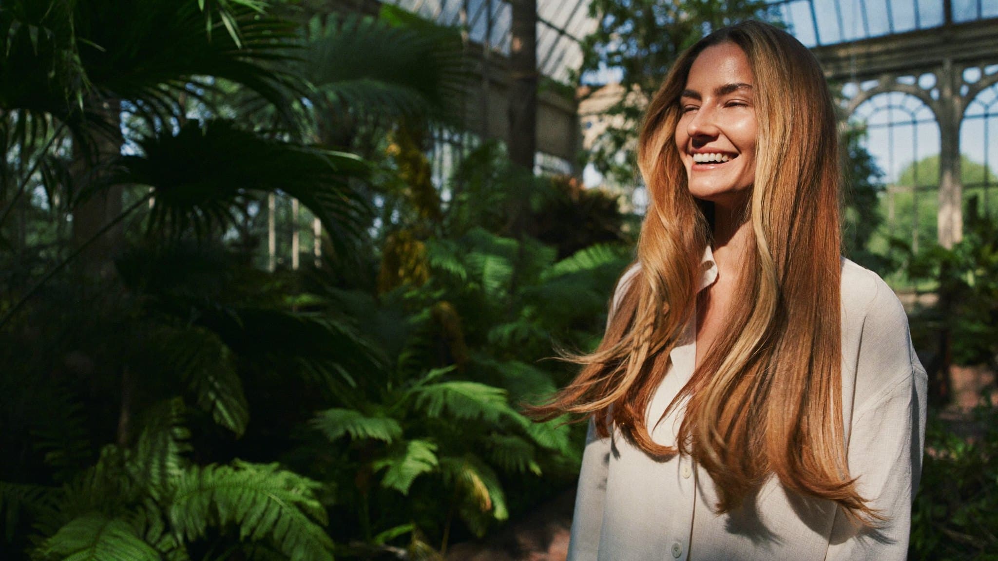 A woman laughing in a sunlit Victorian glasshouse, her long honey-brunette hair swinging.