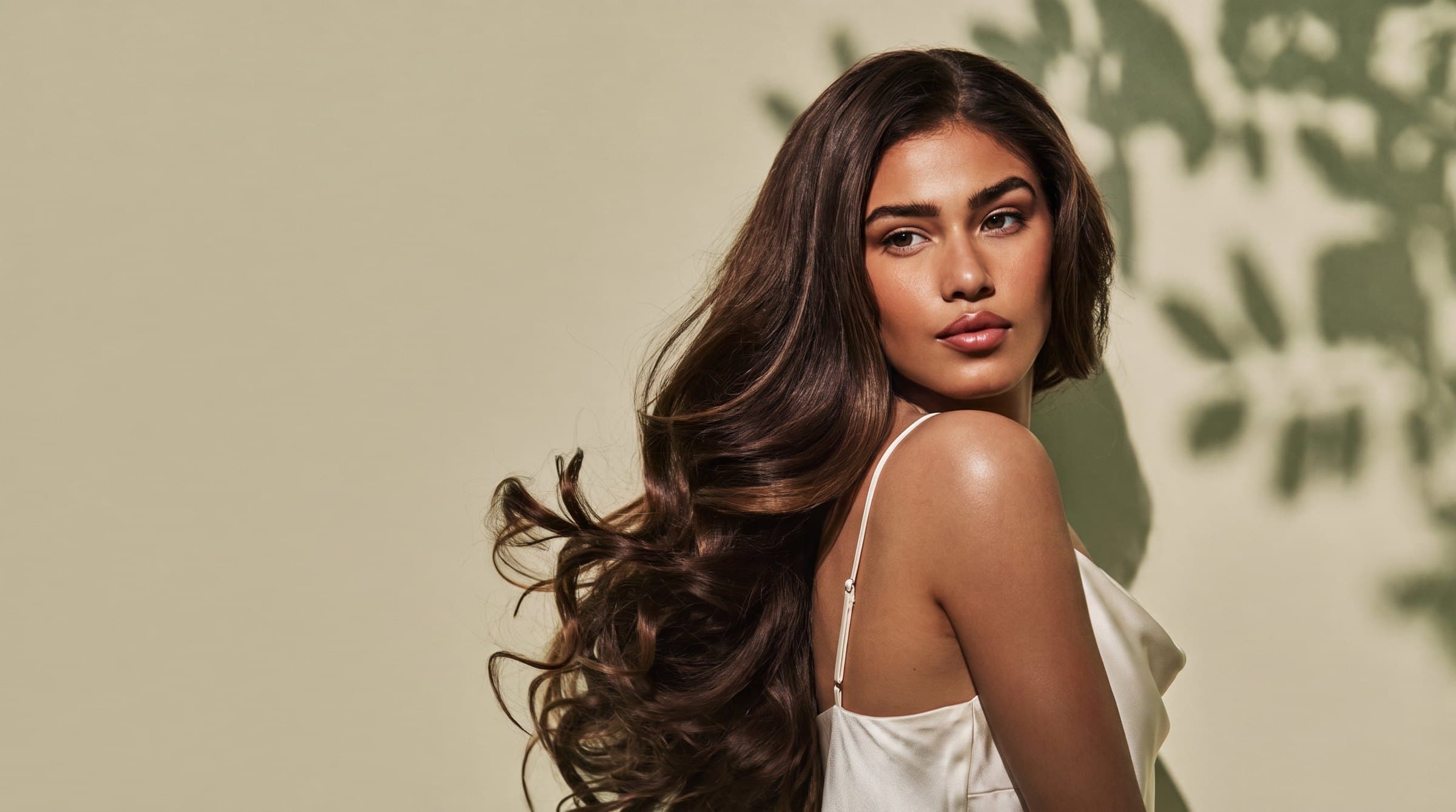 A woman with long, glossy brunette waves in an ivory silk top, glancing over her shoulder against a sunlit sage-green wall dappled with leaf shadows.