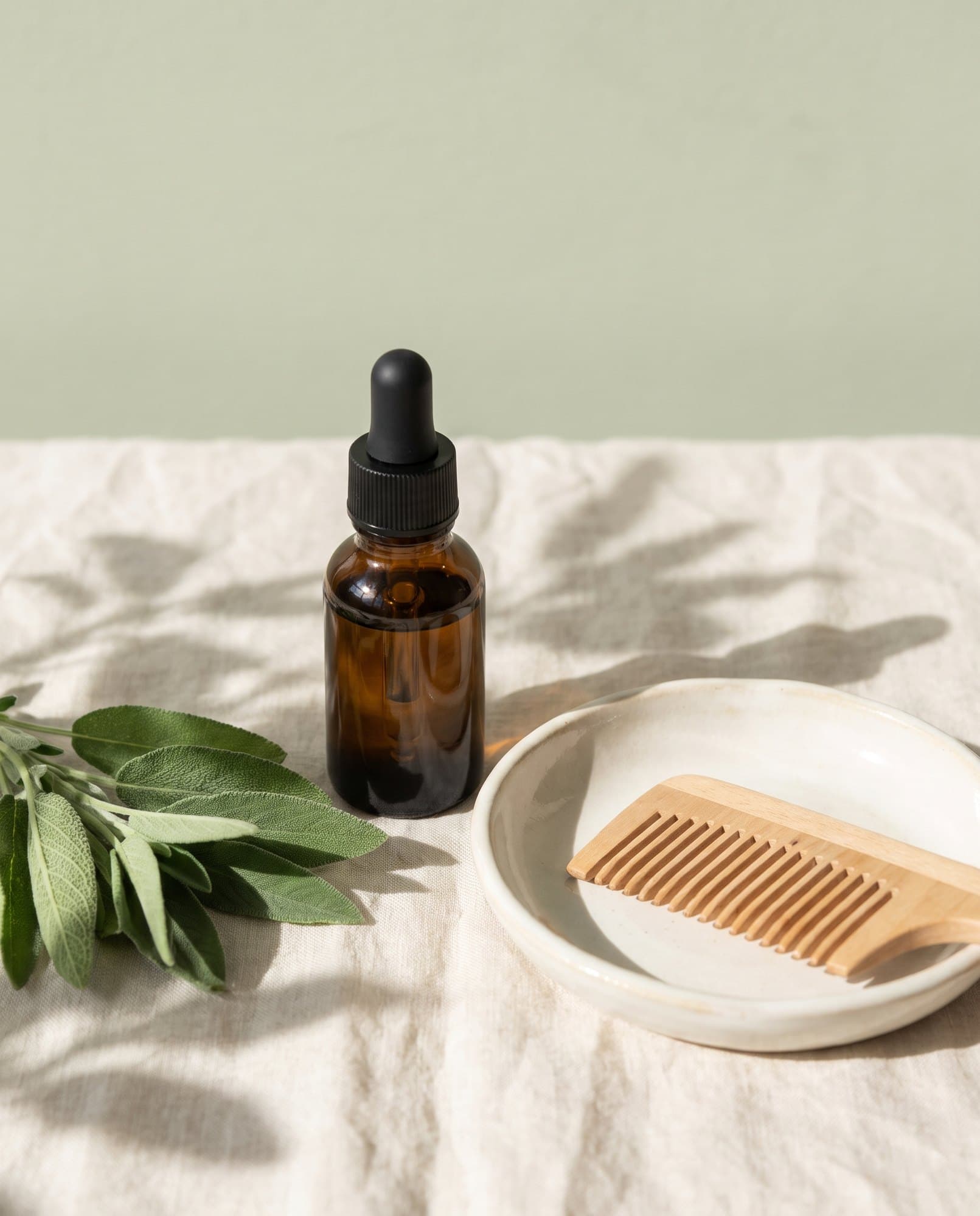 A hair oil dropper bottle, a wooden comb and sage leaves on cream linen.